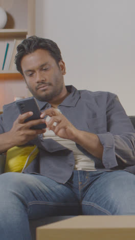 Vertical-Video-Of-Multi-Cultural-Friends-Sitting-On-Sofa-At-Home-Together-All-Looking-At-Their-Mobile-Phones